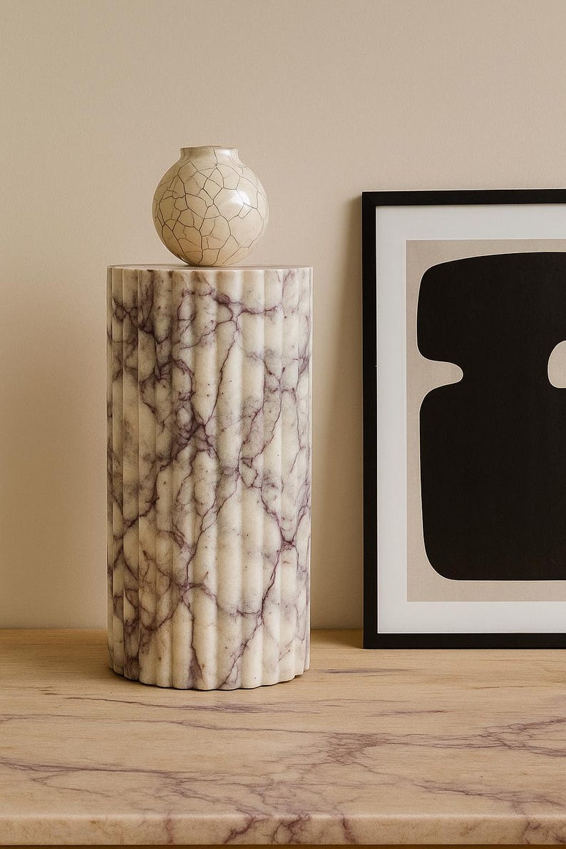 May include: A cylindrical marble pedestal with vertical fluting and a cream and purple veined pattern. A small, round, cream-colored vase with a cracked glaze sits atop the pedestal. A framed abstract art piece is to the right.