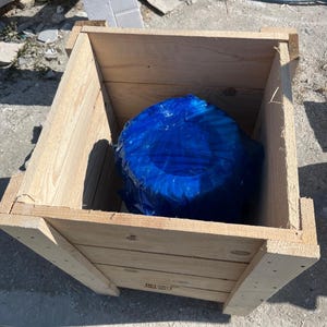 May include: A light-colored wooden crate with a blue object inside. The crate is constructed from planks of wood and has a rectangular shape. The blue object is wrapped in plastic.
