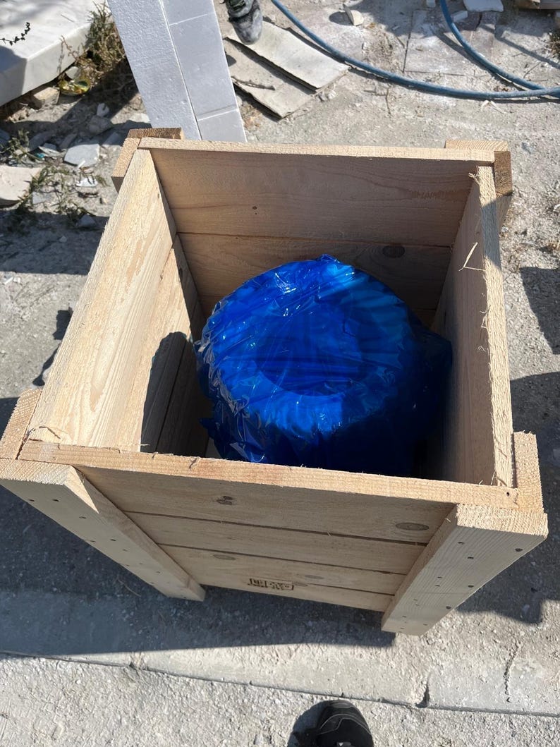 May include: A light-colored wooden crate with a blue object inside. The crate is constructed from planks of wood and has a rectangular shape. The blue object is wrapped in plastic.