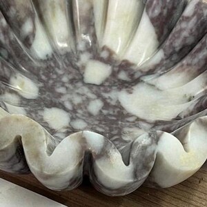 Pu&ograve; includere: Una ciotola decorativa in marmo con bordo smerlato. La superficie della ciotola presenta un motivo naturale di tonalit&agrave; bianche, grigie e viola. I bordi ondulati e l'interno hanno una finitura liscia e lucida.