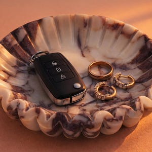 Pu&ograve; includere: Un vassoio decorativo in marmo con bordo smerlato contiene una chiave dell'auto nera, una fede nuziale d'oro e due orecchini a cerchio d'oro con pietre trasparenti. Il vassoio ha un motivo marmorizzato crema e viola.