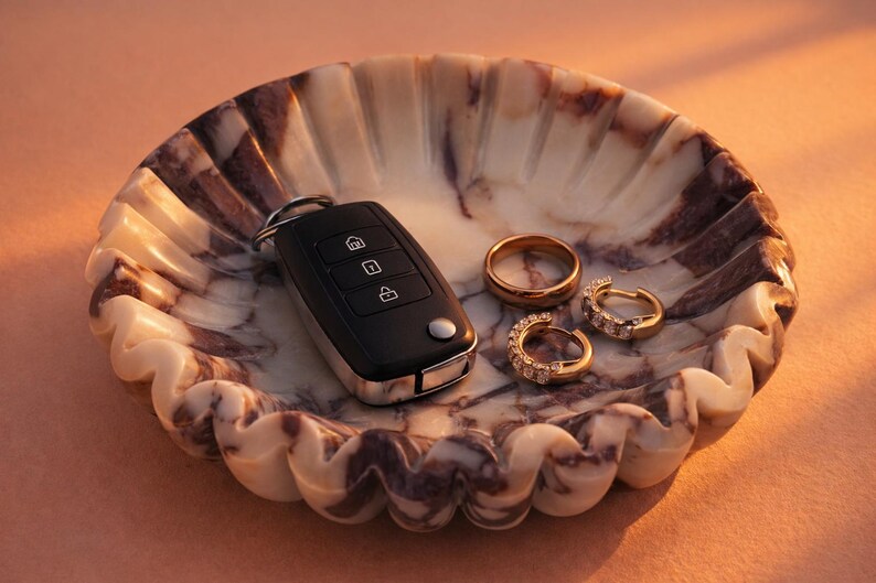 Pu&ograve; includere: Un vassoio decorativo in marmo con bordo smerlato contiene una chiave dell'auto nera, una fede nuziale d'oro e due orecchini a cerchio d'oro con pietre trasparenti. Il vassoio ha un motivo marmorizzato crema e viola.