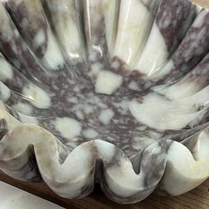 Pu&ograve; includere: Una ciotola decorativa in marmo con bordo smerlato. La ciotola presenta un motivo naturale di venature bianche e grigio-viola. Il design ricorda una conchiglia, con un bordo ondulato e strutturato. Ideale per l'arredamento della casa, circa 25 cm di diametro.