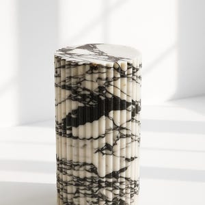 May include: A cylindrical marble side table with a fluted design. The table features a white base with black veining, and a flat, circular top. The design is modern and minimalist, suitable for various interior styles.