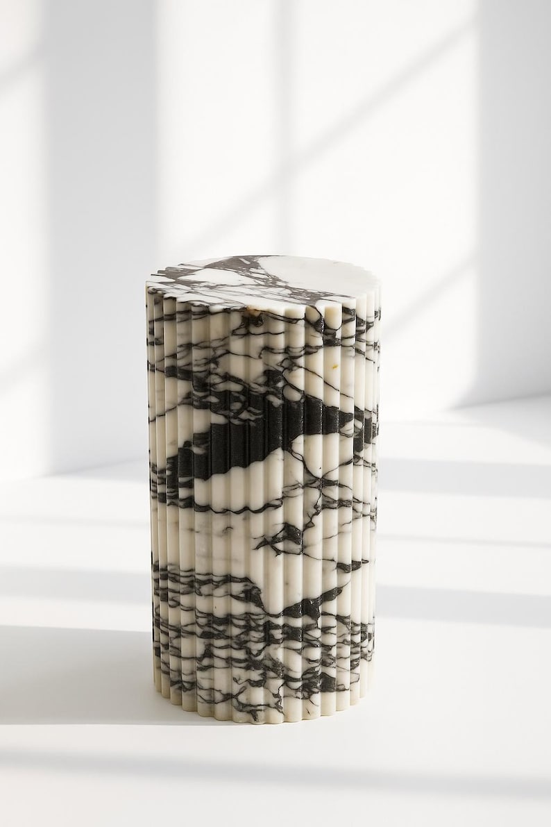 May include: A cylindrical marble side table with a fluted design. The table features a white base with black veining, and a flat, circular top. The design is modern and minimalist, suitable for various interior styles.