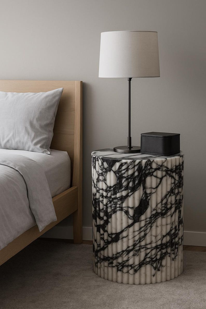 modern bedroom interior featuring a luxurious black and white fluted marble nightstand placed beside a light wood bed frame. The cylindrical marble table displays striking dark veining against a creamy white base, complementing the neutral-toned decor. On top sits a sleek black box and a minimalist lamp with a white shade, creating a serene and elegant aesthetic. Soft natural lighting enhances the stone’s texture, highlighting its craftsmanship and timeless appeal in a contemporary spacete