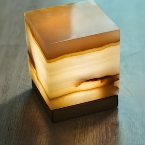 Può includere: Lampada rettangolare in onice, che emana un bagliore caldo. La lampada presenta tonalità ambra e crema, con un design a strati. Poggia su una base in legno marrone scuro. La luce emana dall'interno, creando un'atmosfera soffusa.