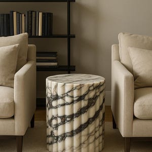 May include: A cylindrical marble side table with a fluted design. The table features a white base with dark gray veining. The table is placed between two beige armchairs in a living room setting.