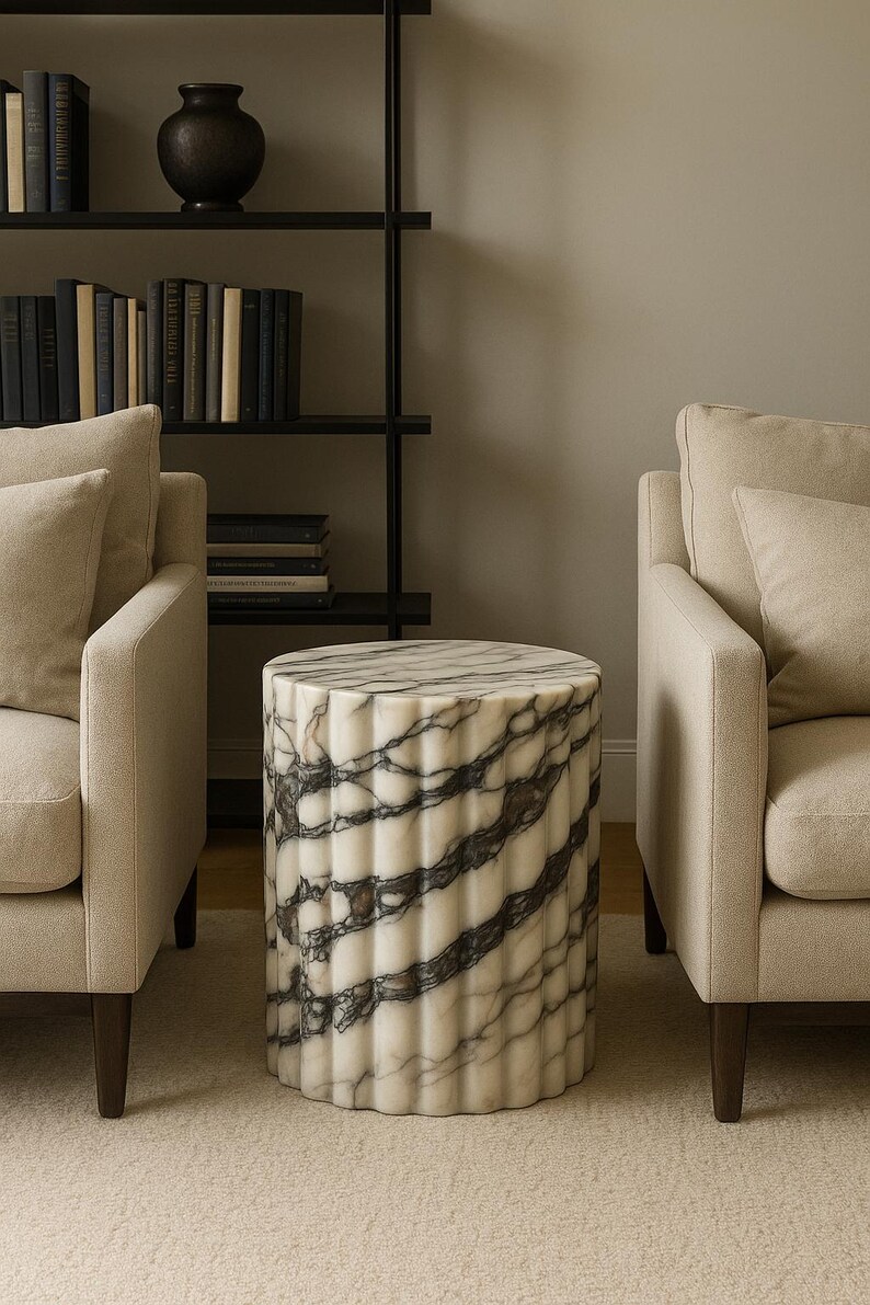 May include: A cylindrical marble side table with a fluted design. The table features a white base with dark gray veining. The table is placed between two beige armchairs in a living room setting.