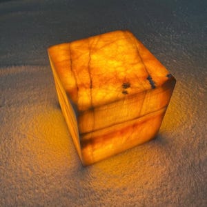 May include: A glowing, amber-colored onyx cube. The translucent stone reveals natural veining and patterns, creating a warm, inviting ambiance. The cube is illuminated from within, showcasing its unique texture and color variations.