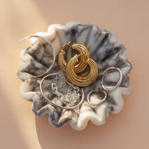 Pu&ograve; includere: Una collezione di orecchini e bracciali in oro e argento esposti in una ciotola decorativa in marmo. Gli orecchini includono grandi orecchini a cerchio dorati, piccoli orecchini a cerchio argentati e un braccialetto a catena d'argento. La ciotola ha un motivo marmorizzato bianco e grigio.