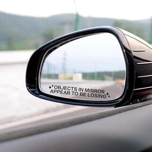 May include: A black car side mirror with the text "OBJECTS IN MIRROR APPEAR TO BE LOSING" in white. The mirror reflects a blurred outdoor scene, with a focus on the road and the surrounding environment.