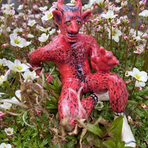 May include: A red ceramic figurine of a devil with horns and a black spotted pattern, sitting in a bed of white flowers.