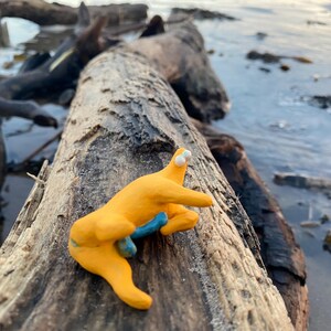 May include: A whimsical, handmade yellow clay figure with blue accents, posed on a weathered log at the beach. The figure has two white eyes and outstretched arms. The background shows water and other logs.