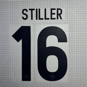 Peut inclure: Vinyle thermocollant noir avec le nom "STILLER" au-dessus du numéro "16". Le numéro est grand et en gras. Le vinyle est sur une feuille de support transparente avec un motif quadrillé.
