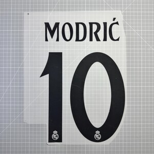 Peut inclure: Panneau blanc avec le nom "MODRIĆ" en lettres noires au-dessus du numéro "10". Le numéro est également en noir. De petits écussons sont en bas. Le panneau est sur un fond de grille grise.