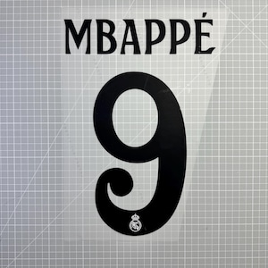 Peut inclure: Vinyle thermocollant noir avec le nom "MBAPPÉ" au-dessus du chiffre "9". Le chiffre a un ornement décoratif et un petit emblème en bas. Le vinyle est sur un support transparent.