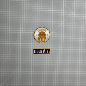Peut inclure: Un écusson brodé circulaire doré et blanc avec le texte CHAMPIONS 2025 et un graphique central, accompagné d'un écusson rectangulaire noir avec le texte blanc et doré LIGUE 1 M, tous deux conçus pour être appliqués sur du tissu.