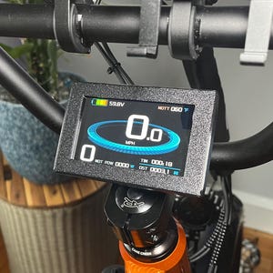 May include: Close-up of an electric bike's digital display showing a speed of 0.0 MPH. The screen displays battery voltage, temperature, and other metrics. The bike's handlebars and frame are visible in the background.