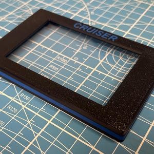 May include: Black rectangular frame with the word "CRUISER" in blue lettering. The frame has a textured surface and a thin blue stripe along one edge. The frame is on a blue cutting mat.