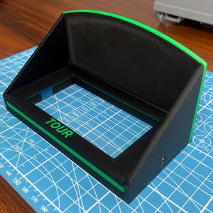 May include: A black monitor hood with a bright green trim and the word TOUR printed in green on the front frame. The device features a rectangular opening and a slanted top panel designed to reduce glare on a screen, with a matte finish.