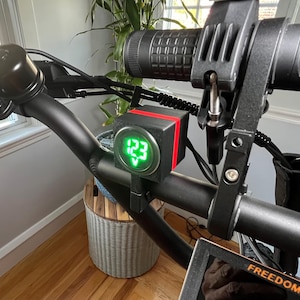 May include: Close-up of a black electric scooter handlebar with a digital display showing "123" in green. The display is mounted on a black cube with a red stripe. The scooter has a black handlebar and a black control panel.
