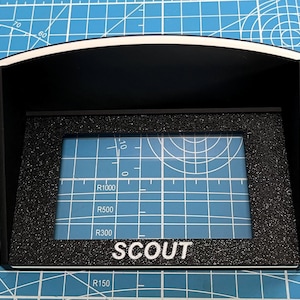 May include: A black and white electronic device with the word "SCOUT" printed on the bottom. The device has a screen with grid lines and measurement markings. The device is on a blue surface.