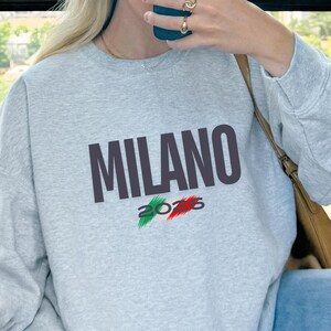 Puede incluir: Sudadera gris ceniza con la palabra "MILANO" en letras oscuras. Debajo, el número "2026" con los colores de la bandera italiana. La sudadera es de cuello redondo.