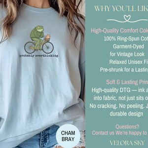 May include: Light blue t-shirt featuring a green frog riding a bicycle with a basket. The frog is wearing a hat and the text "probably overthinking" is below the graphic. The shirt is made of 100% ring-spun cotton.