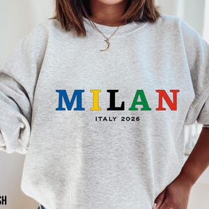Puede incluir: Sudadera gris claro con la palabra "MILAN" en letras azules, amarillas, negras y rojas. Debajo, el texto "ITALY 2026" está impreso en negro. La sudadera es de cuello redondo.
