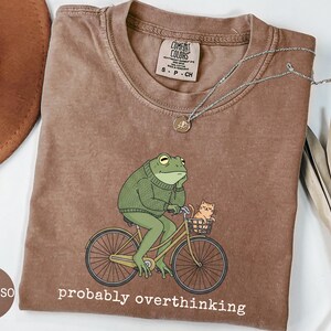May include: Espresso-colored t-shirt featuring a cartoon frog riding a bicycle with a cat in the basket. The frog wears a green sweater and the text "probably overthinking" is printed below the image. A silver necklace is draped across the shirt.