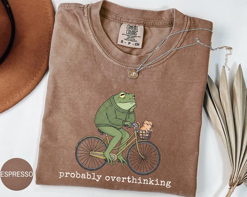 May include: Espresso-colored t-shirt featuring a cartoon frog riding a bicycle with a cat in the basket. The frog wears a green sweater and the text "probably overthinking" is printed below the image. A silver necklace is draped across the shirt.