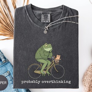 May include: Dark gray Comfort Colors t-shirt featuring a green frog riding a yellow bicycle with a small orange cat in the basket. The text "probably overthinking" is printed below the graphic. A silver necklace is draped over the shirt.