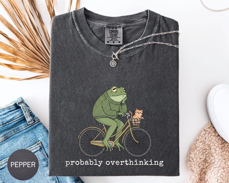 May include: Dark gray Comfort Colors t-shirt featuring a green frog riding a yellow bicycle with a small orange cat in the basket. The text "probably overthinking" is printed below the graphic. A silver necklace is draped over the shirt.