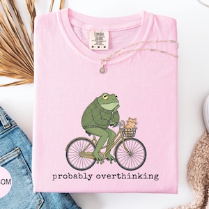 May include: A light pink t-shirt with a graphic of a green frog riding a bicycle with a cat in the basket. The text "probably overthinking" is below the graphic. The shirt has a Comfort Colors tag.