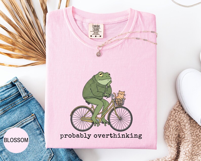 May include: A light pink t-shirt with a graphic of a green frog riding a bicycle with a cat in the basket. The text "probably overthinking" is below the graphic. The shirt has a Comfort Colors tag.