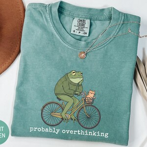 May include: Light green t-shirt with a graphic of a frog riding a bicycle with a cat in the basket. The frog wears a green sweater and is deep in thought. The text "probably overthinking" is printed below the graphic.