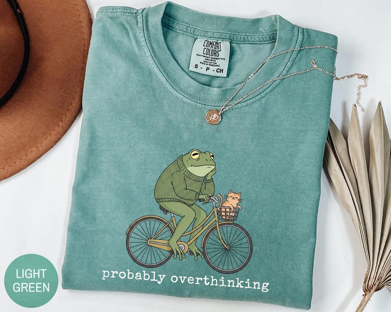 May include: Light green t-shirt with a graphic of a frog riding a bicycle with a cat in the basket. The frog wears a green sweater and is deep in thought. The text "probably overthinking" is printed below the graphic.