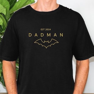 Peut inclure: T-shirt noir avec texte doré et symbole de chauve-souris. Le texte indique "EST 2024 DADMAN" avec une silhouette de chauve-souris en dessous. Le t-shirt est de couleur noire unie.