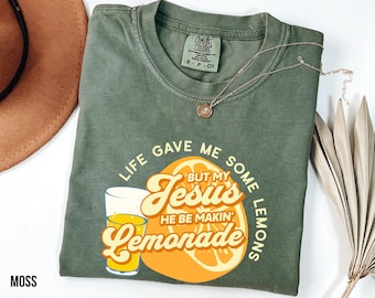 May include: Moss green t-shirt with the text "Life gave me some lemons but my Jesus he be makin' lemonade" in yellow and orange. The shirt features a graphic of a glass of lemonade and a lemon slice. A gold necklace is draped across the shirt.