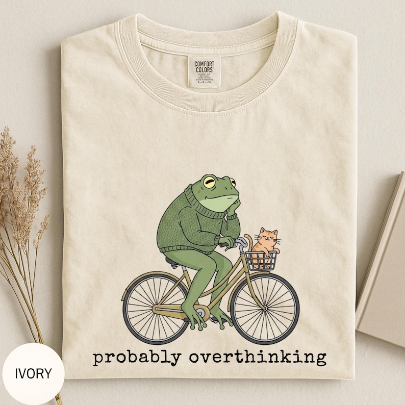 May include: Ivory-colored t-shirt featuring a cartoon frog riding a bicycle with a cat in the basket. The frog wears a green sweater and appears to be deep in thought. The text "probably overthinking" is printed below the image.