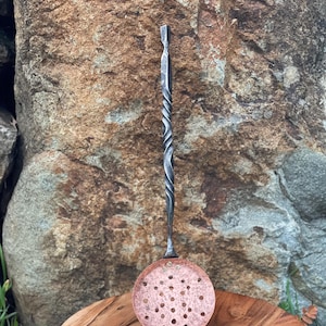 May include: A hand-forged metal skimmer with a copper-colored, perforated round head and a twisted handle. The skimmer is displayed on a wooden surface against a natural stone backdrop. This kitchen utensil is designed for straining foods.
