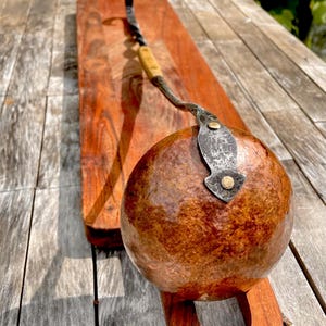 May include: A handcrafted copper and metal fire pit poker with a wooden handle, resting on a wooden cutting board. The poker is designed with a curved handle and a decorative metal end. The fire pit poker is a rustic, functional item.