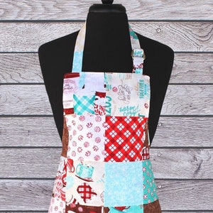 May include: A patchwork kitchen apron with a red, white, and blue color scheme. The apron features a variety of patterns, including plaid, polka dots, and floral prints. The apron has a brown and white text pattern that says "Cozy" and "Coffee".