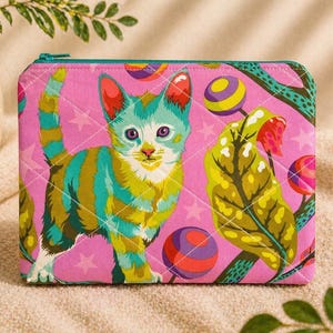 May include: A quilted pink zippered pouch featuring a colorful cat illustration with teal and yellow stripes. The design includes whimsical elements like stars, lollipops, and stylized leaves. The pouch has a teal zipper.