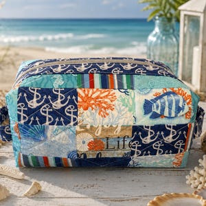 May include: A quilted zippered pouch featuring a patchwork design with nautical themes including navy blue anchors, orange coral, striped patterns, and a blue fish. The fabric includes the text Enjoy Life in a script font on a light background.