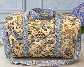 May include: A quilted duffel bag featuring a cream-colored fabric printed with a pattern of various teapots, teacups, and floral arrangements. The bag includes two top handles and a long shoulder strap, all covered in a blue floral print.