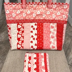 May include: A pink tote bag and matching wallet sit on a chair. The tote bag has pink heart patterns and red handles. The wallet and the bottom of the tote bag have vertical stripes of red and pink fabrics.