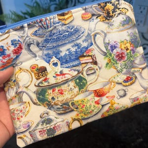 May include: A quilted pouch with a blue zipper, featuring a repeating pattern of teacups, teapots, and pastries. The design includes floral and ornate details in shades of blue, green, yellow, and red on a cream background.