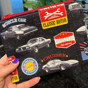 May include: Black zippered pouch with a red zipper and trim, featuring a vintage car theme. The design includes classic car illustrations, the words "MUSCLE CAR", "CLASSIC MOTOR", "CRUISER", and "VINTAGE GARAGE".